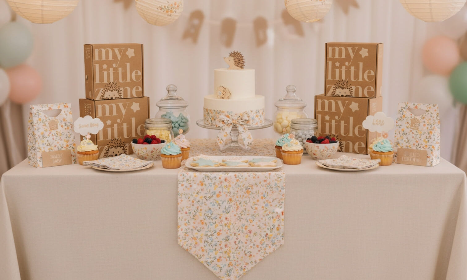Baby shower dessert table setup with a floral theme, featuring a decorated cake, cupcakes, pastel balloons, and gift boxes arranged for a baby celebration.