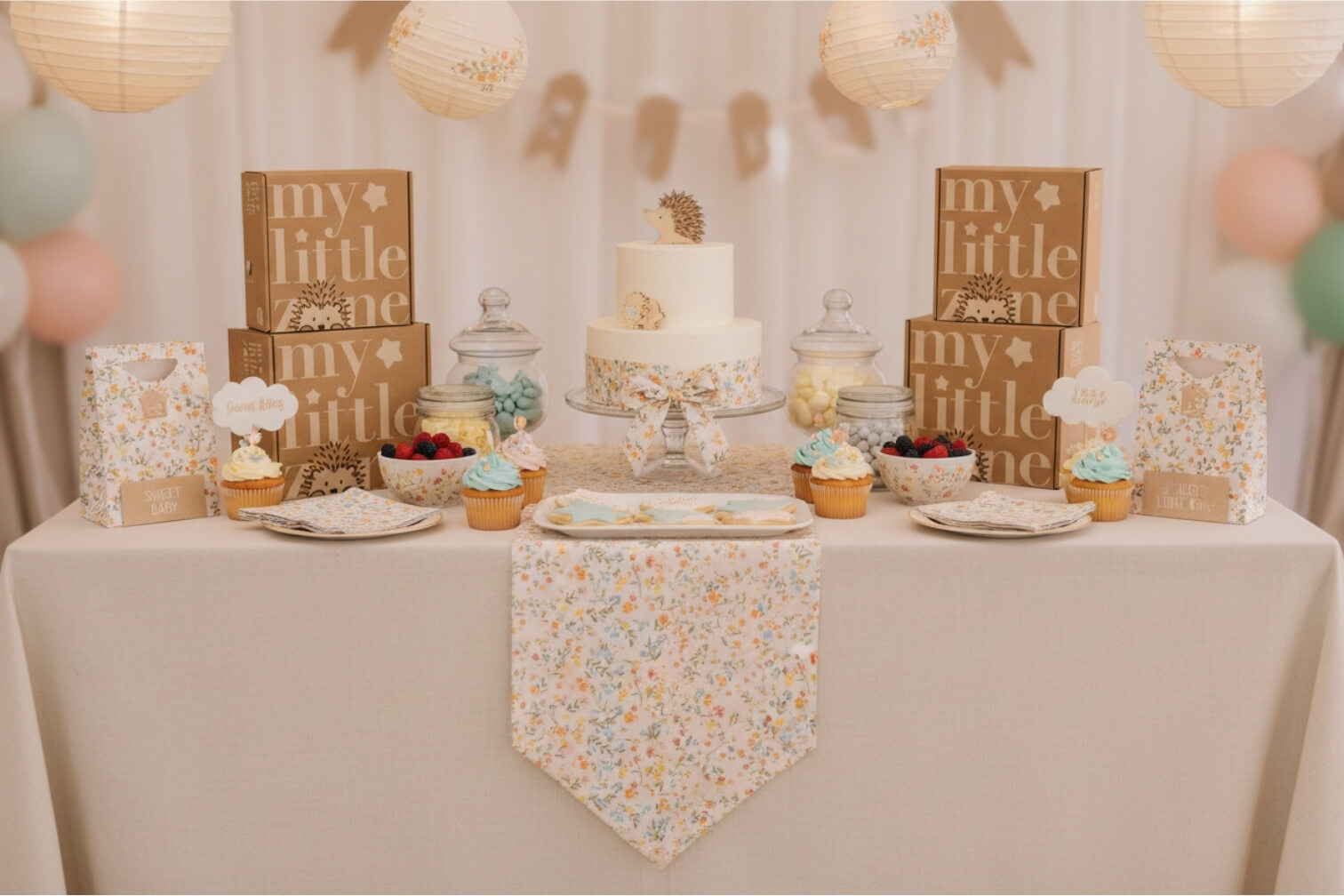 Baby shower dessert table setup with a floral theme, featuring a decorated cake, cupcakes, pastel balloons, and gift boxes arranged for a baby celebration.
