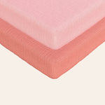 Solid coral and solid pink muslin bassinet sheets on a bed with a white background