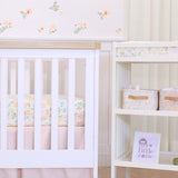 Floral-themed baby nursery with a white crib, floral patterned muslin crib bedding and changing pad station with storage bins on it's shelves.