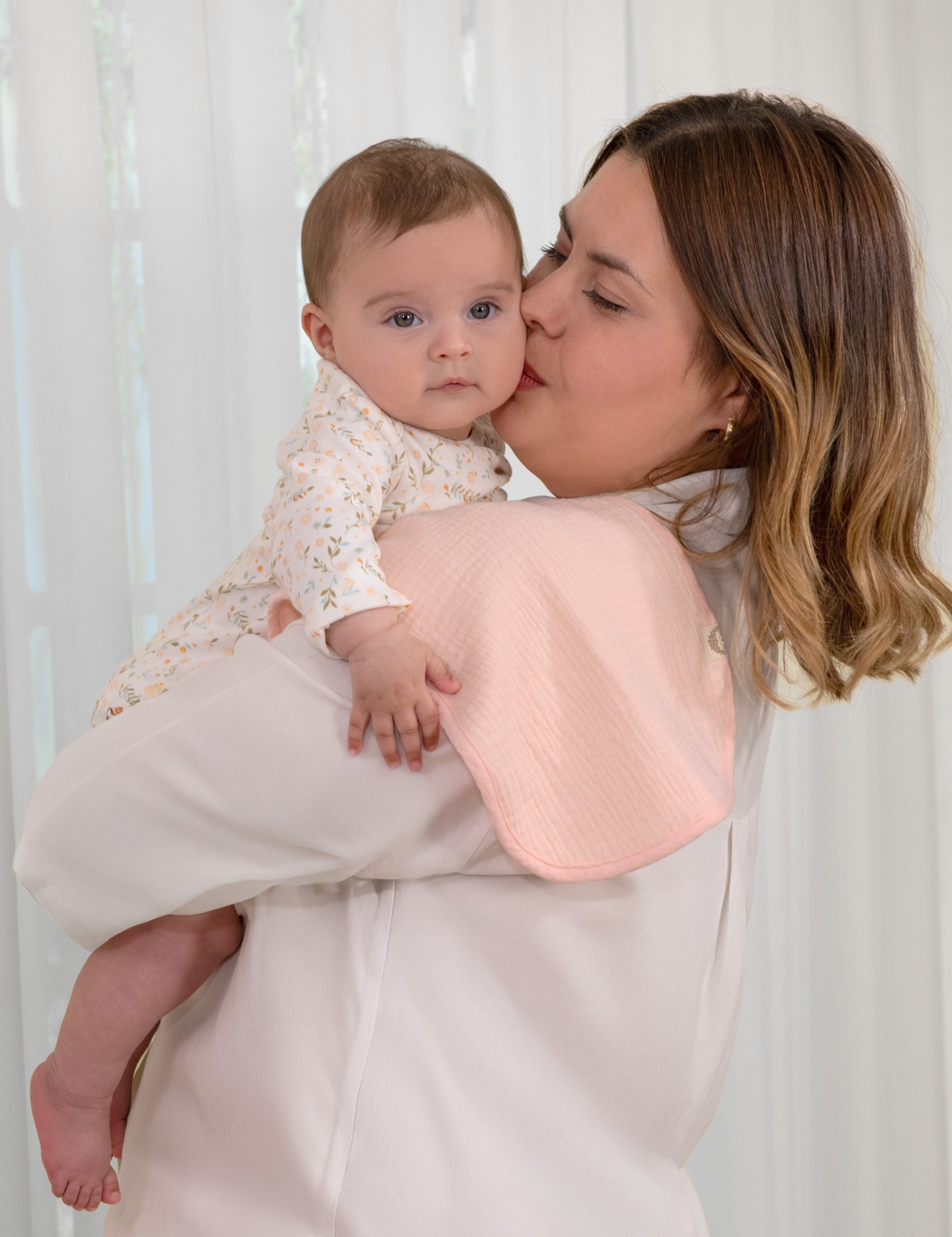 Mother holding her baby and kissing it on the cheek and carrying solid pink muslin burp cloths on her shoulder.