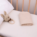 Baby crib with a plush bunny toy and folded solid beige bassinet sheet on a white muslin crib fitted sheet.
