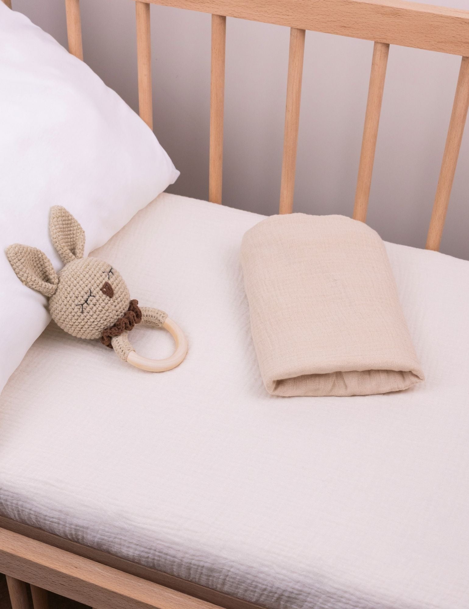 Baby crib with a plush bunny toy and folded solid beige bassinet sheet on a white muslin crib fitted sheet.