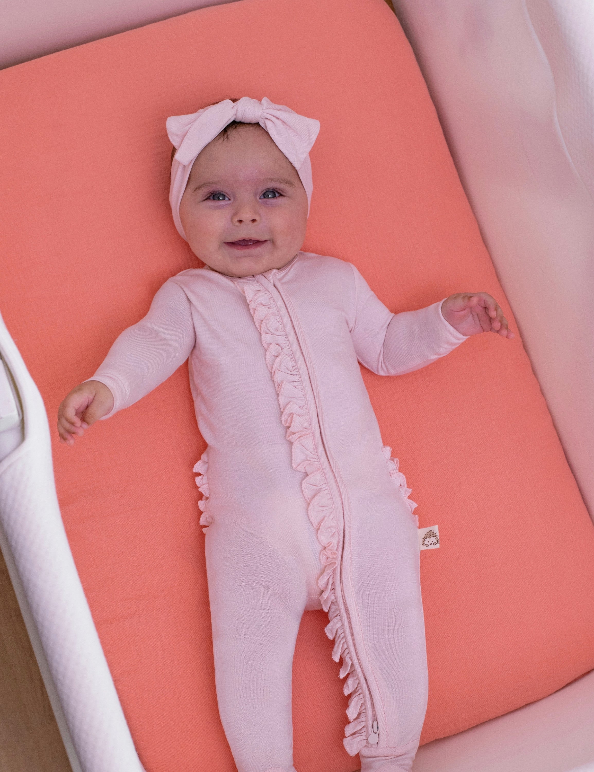 Baby in a solid pink ruffled zipper and headband lying on a solid coral muslin bassinet sheet.