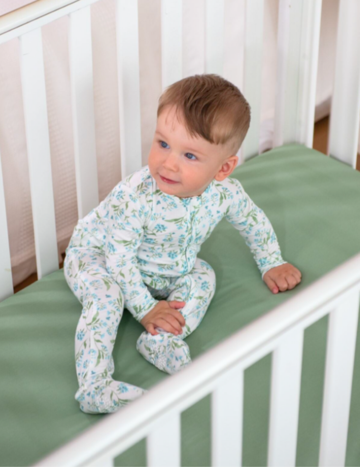 Baby in a floral onesie sitting on a crib with green fitted mattress sheet.