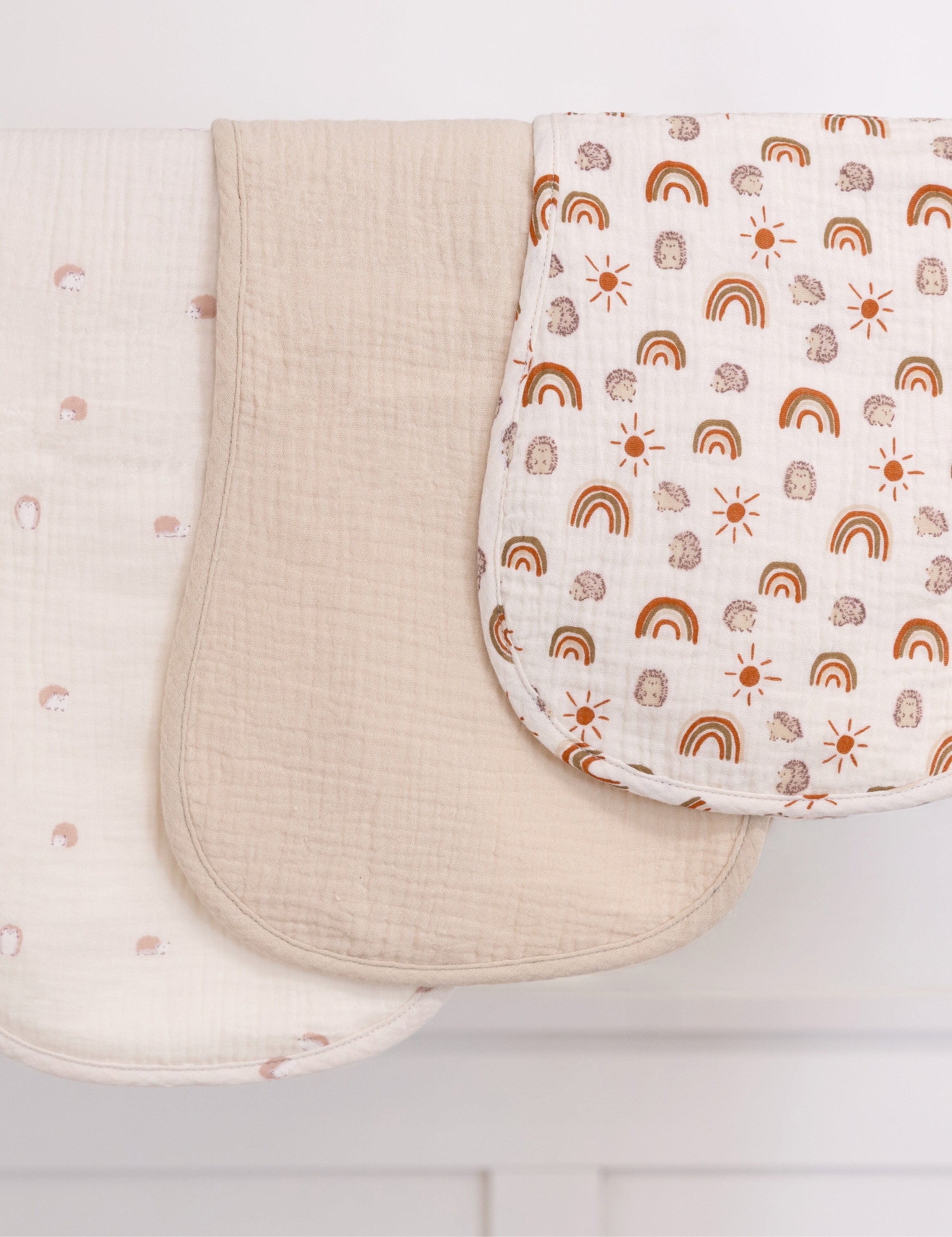 Three burp cloths, one solid beige, one with rainbow pattern and one with hedgehog illustrations.
